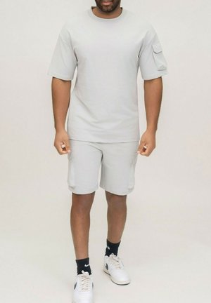 Homme debout portant une chemise à manches courtes gris clair et un short cargo assorti, avec des chaussettes noires et des baskets Nike blanches