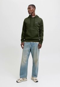 Olijfgroene hoodie met een kangoeroezak en verstelbare trekkoord capuchon. Draagt een lichtblauwe spijkerbroek en beige sneakers.