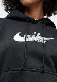 Person iført en svart hettegenser med hvit, lodden Nike-logo og snorer, som viser nedre del av ansiktet og krøllete hår.