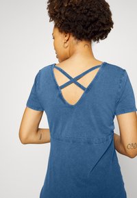 Vestido de ganga azul com mangas curtas e costas em V pronunciado, apresentando alças cruzadas. Textura suave com um corte descontraído e detalhe de costura na cintura.