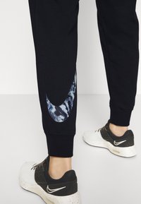 Svarta sweatpants med en stor reflekterande Nike-logga i ett vågmönster på sidan, tillsammans med svarta och vita sneakers.