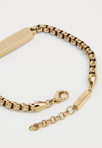 Bracelet en chaîne dorée avec des maillons rectangulaires et ronds, un fermoir mousqueton, et une étiquette décorative avec un logo gravé. La texture de la chaîne est lisse et brillante.