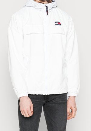 Homme portant une veste à capuche blanche Tommy Jeans avec des poignets élastiques et un jean noir, debout devant un fond clair uni.