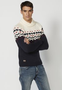 Suéter de punto con un cuello alto, sección superior color crema y mitad inferior color navy. Presenta un patrón geométrico en rojo y negro en el pecho.