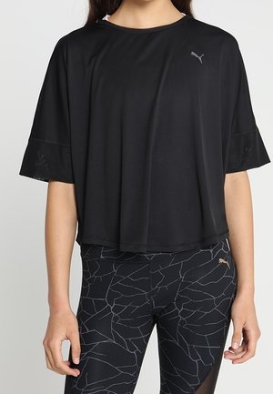 Kobieta ubrana w luźną czarną koszulkę Puma i czarne legginsy z wzorem oraz logo Puma, stojąca z ramionami swobodnie opuszczonymi wzdłuż ciała.