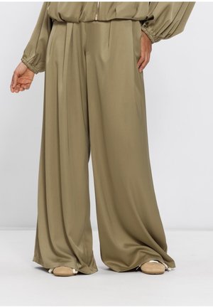 Wijde kaki broek met plooien, gedragen met een bijpassend jasje en beige sneakers, waarop de onderkant van de romp en benen te zien zijn.