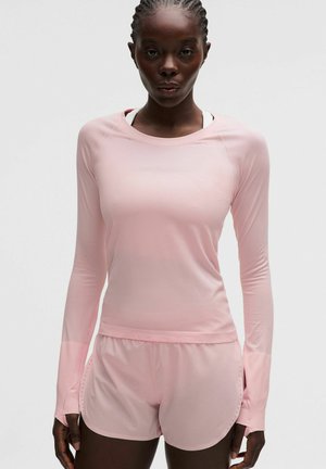 Mujer con el cabello trenzado, vestida con una camisa de manga larga color rosa claro y pantalones cortos a juego, de pie frente a un fondo blanco liso.