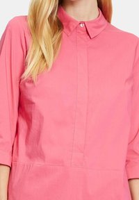 Camicia rosa con colletto e maniche tre quarti, realizzata in tessuto liscio e dotata di patta con bottoni e vita arricciata per una maggiore ampiezza.