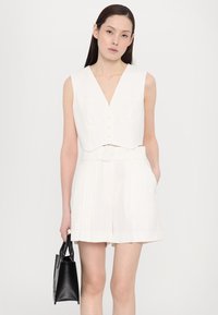 Chaleco corto blanco con escote en V y cierre de botones, combinado con pantalones cortos de rayas verticales de talle alto y un pequeño bolso negro.