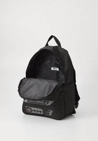 Sac à dos noir fabriqué en matériau durable, avec une ouverture zippée, un intérieur doublé et un logo gris bien visible sur le devant.