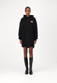 Vestido hoodie oversized preto com um bolso frontal e logo "DIESEL" em vermelho. Apresenta capuz e mangas longas, combinado com botas pretas até ao joelho.