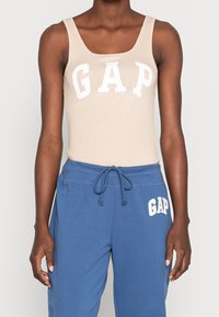 Débardeur beige à côtes avec un grand logo blanc "GAP", associé à un pantalon de jogging bleu avec un plus petit logo blanc "GAP" sur la jambe.
