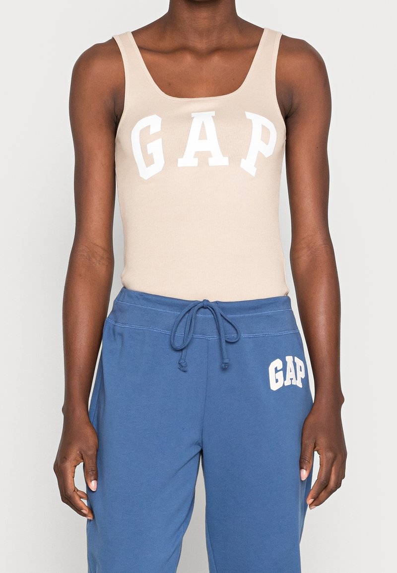 Débardeur beige à côtes avec un grand logo blanc "GAP", associé à un pantalon de jogging bleu avec un plus petit logo blanc "GAP" sur la jambe.