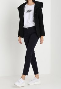 Schwarze Kapuzenjacke über einem weißen T-Shirt mit schwarzem Druck, kombiniert mit marineblauen Pants und weißen Sneakern. Lässiges Outfit mit moderner Silhouette.