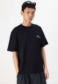 Tricou din bumbac negru, cu o croială lejeră, guler rotund și mâneci scurte; prezintă cuvântul "Premium" brodat în alb pe pieptul stâng.