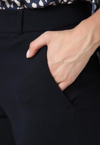 Main avec un vernis à ongles rose clair reposant dans la poche avant d’un pantalon bleu marine foncé, avec un haut noir et crème à motifs visible.