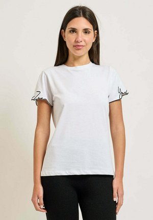 Giovane donna con lunghi capelli scuri che indossa una maglietta bianca a maniche corte con bordi neri scritti sulle maniche e pantaloni neri, in piedi.