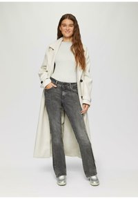 Hellgrauer Übermantel, gerippter cremefarbener Pullover und dunkelgraue Jeans. Die Sneakers sind metallic silber. Das Modell steht aufrecht mit einer Hand in der Jeans-Tasche.