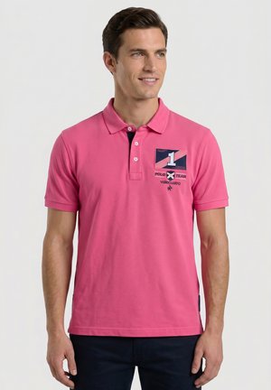 Hombre con una camiseta polo de color rosa brillante con un logo parche en el pecho izquierdo, de pie contra un fondo liso y claro.