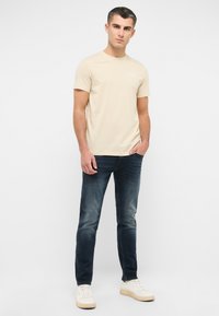 Camiseta de manga corta color beige claro con cuello redondo, combinada con jeans azul oscuro y zapatillas blancas. La tela parece suave y casual.
