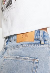 Světle modré džínové kalhoty mají zadní kapsu s opotřebovaným detailem a hnědou koženou nášivku označenou "BDG" na pasu.