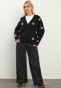 Czarny sweter kardigan z białym haftem kwiatowym, dekoltem w serek i zapięciem na guziki. W zestawie z szerokimi czarnymi spodniami i trampkami.