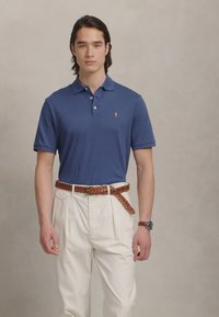 Temno modra polo majica s tremi gumbi, oblečena z bledo kremnimi nabranimi hlačami in tkanim rjavim usnjenim pasom. Ura na levi zapestju.