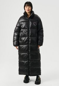 Mujer de pie con un largo abrigo acolchado negro con cremallera frontal y un logo de corazón rosa en el pecho, y zapatos de plataforma negros.