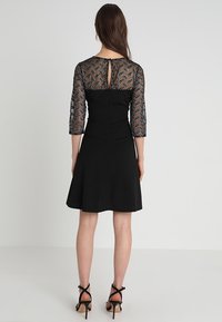 Svart klänning med trekvarts genomskinliga mönstrade ärmar, figursydd överdel, utsvängd kjol och en nyckelhålsdetalj på ryggen. Bärs med svart klackskor med remmar.