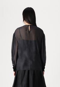 Zwarte, doorschijnende blouse met lange mouwen, voorzien van een knoopsluiting bij de manchetten en een keyhole-detail aan de achterkant, gecombineerd met een zwarte rok.