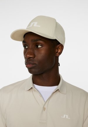 Beige Kappe mit einem flachen Schild und einem Logo auf der Vorderseite. Die Kappe hat eine glatte Textur. Das Modell trägt ein passendes beige Poloshirt mit einem weißen Unterhemd.