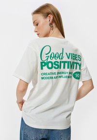Hvid bomulds T-shirt med grøn trykt tekst p�å ryggen: "Good Vibes Positivity" med yderligere sætninger. Løs pasform og korte ærmer.