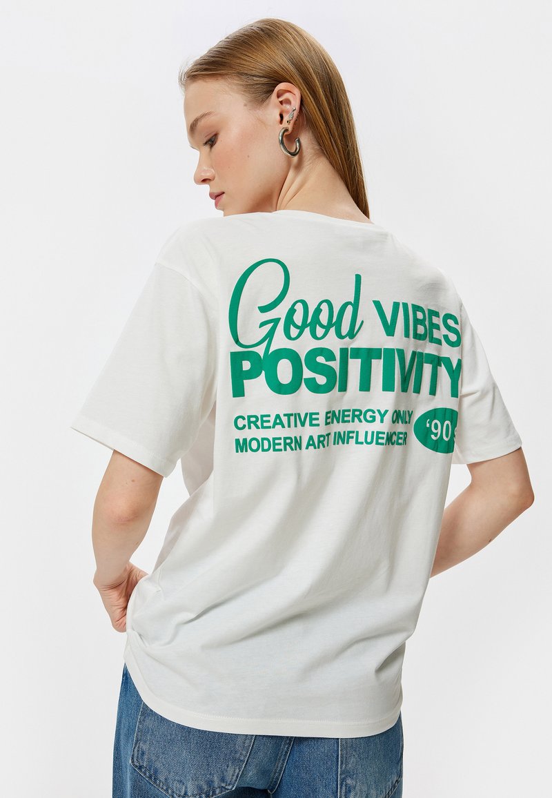 Hvid bomulds T-shirt med grøn trykt tekst på ryggen: "Good Vibes Positivity" med yderligere sætninger. Løs pasform og korte ærmer.