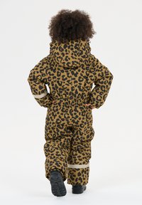 Barn med krøllet hår iført en snowsuit i brunt og sort leopardprint med reflekterende striber, stående med hænderne på hofterne og vendt væk.