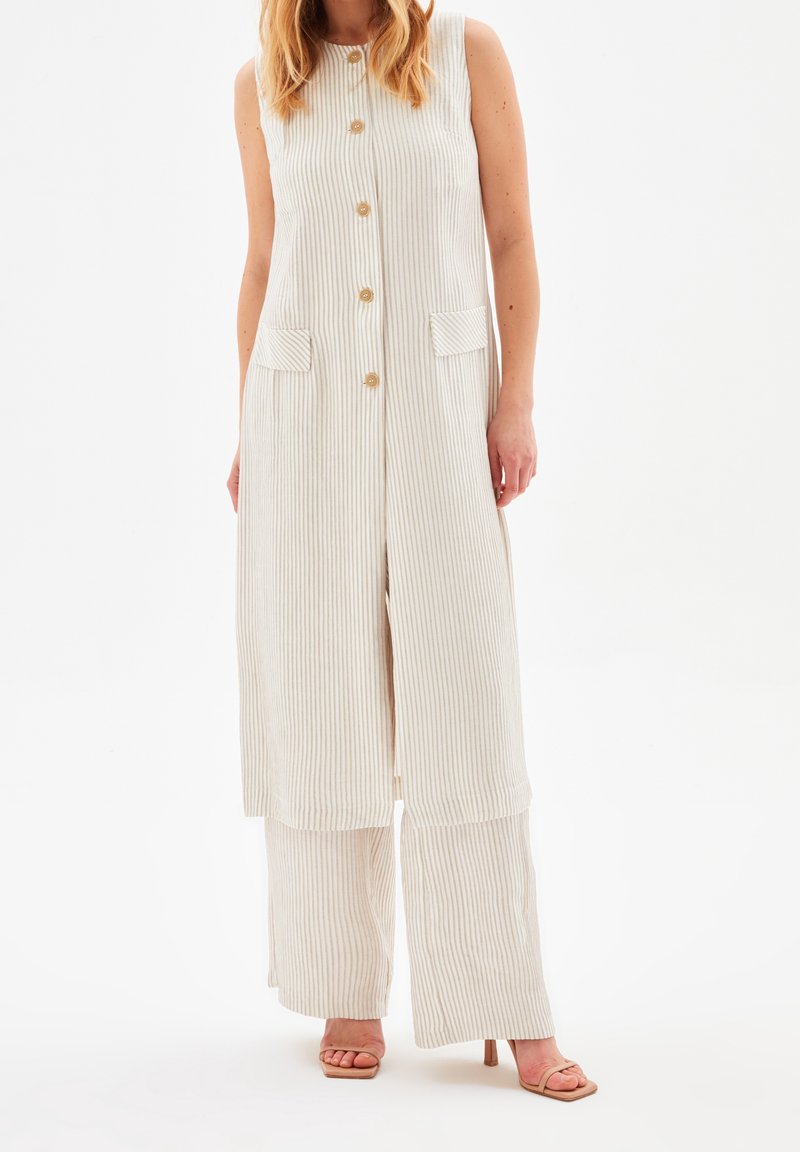 Femme portant un gilet sans manches long beige rayé à boutons, sur un pantalon large assorti et des sandales à talons ouvertes.