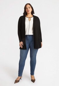 Cárdigan negro sobre una blusa blanca con un corte en forma de llave, combinado con jeans azules y tacones de estampado de leopardo. Presenta un corte relajado y bolsillos frontales.