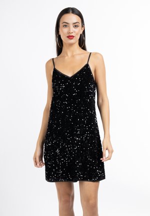 Donna che indossa un abito nero senza maniche con paillettes, spalline sottili e orecchini a cerchio, che posa su uno sfondo bianco semplice.