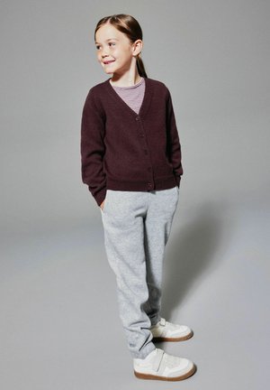 Cardigan borgogna con chiusura a bottoni, abbinato a pantaloni della tuta grigi. Il modello indossa sneaker bianche con dettagli marroni. Camicia a righe semplice sotto.