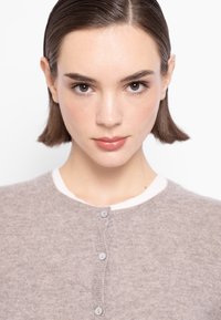 Svetr s kulatým výstřihem v světle hnědé pletenině s bílým límcem. Obsahuje pět šedých knoflíků na přední části. Měkká textura a detailní prošívání.