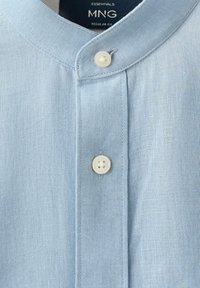 Chemise en lin bleu clair avec col mandarin, deux boutons crème visibles et étiquette MNG Essentials regular fit à l’intérieur du col.