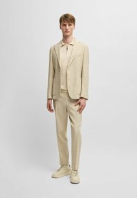 Ljusbeige kostym med texturerad väv, snittlappar och två framfickor. Kombinerad med en beige zip-up polo och krämfärgade sneakers.