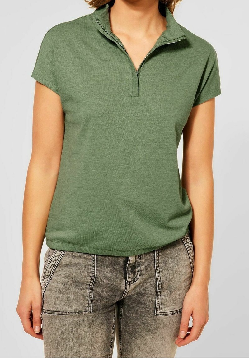 Grön kortärmad polo med dragkedja, tillverkad av mjukt tyg. Matchad med ljusgrå jeans med detaljerad sömnad och fickor.