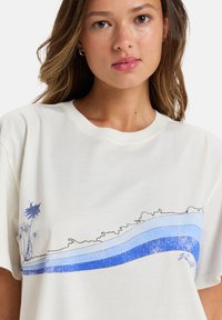 Krämfärgad t-shirt i bomull med blå grafiska linjer och palmträd illustrationer. Avslappnad passform och rund halsdesign.