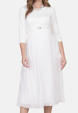 Femme portant une robe midi blanche à manches trois-quarts, jupe plissée, ceinture à la taille et un collier floral distinctif.