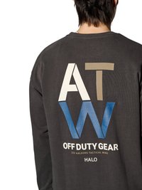 Person iført en mørk langærmet skjorte med store bogstaver "ATW" og teksten "OFF DUTY GEAR 223 AALBORG TACTICAL WING HALO" på ryggen.