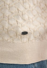 Pull beige clair en maille avec un motif en diamants texturé, doté d'un ourlet côtelé et d'une petite étiquette logo noire.