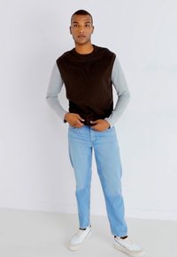 Top senza maniche marrone sopra una maglietta a maniche lunghe grigia chiara, abbinato a jeans blu chiaro e sneakers bianche, in piedi contro uno sfondo bianco.