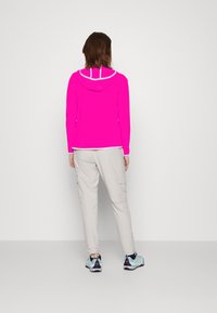 Giacca a vento rosa brillante con accenti bianchi, abbinata a pantaloni cargo beige chiaro. Le scarpe sono di colore blu acqua con dettagli navy e rosa.