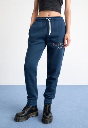 Personne portant un pantalon de survêtement Nike bleu marine avec cordon blanc et des bottes à plateforme noires, debout sur un tapis bleu.