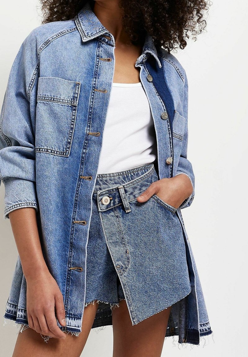 Kvinna som bär en överdimensionerad ljusblå denimjacka, vit linne och en asymmetrisk denimkjol med rå nederkant, med händerna i fickorna.